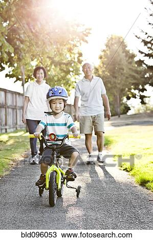 Stock Image - Chinese grandparents watching grandson riding bicycle. Fotosearch