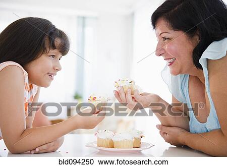 Stock Image - Hispanic mother and daughter eating cupcakes. Fotosearch