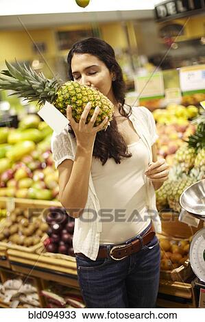Stock Image - Hispanic woman smelling pineapple in grocery store. Fotosearch