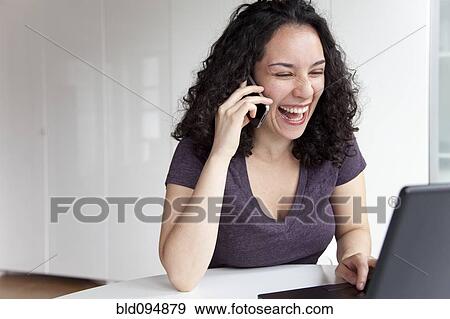 Stock Photo - Hispanic woman talking on cell phone and using laptop. Fotosearch