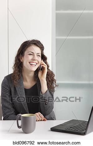 Stock Image - Businesswoman using cell phone at desk. Fotosearch
