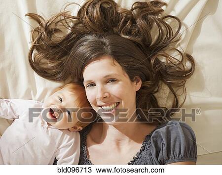 Stock Image - Caucasian mother laying on bed with baby daughter. Fotosearch
