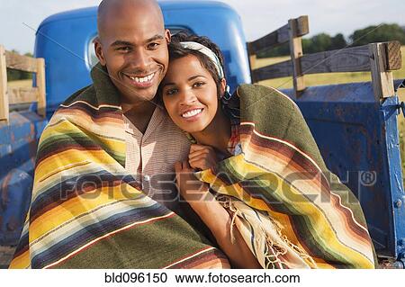 Stock Image - Couple wrapped in blanket sitting on back of truck. Fotosearch