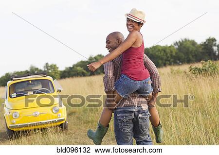 Stock Image - Man piggybacking wife in field near car. Fotosearch