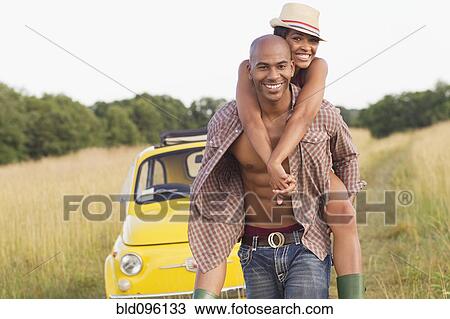 Stock Image - Man piggybacking wife in field near car. Fotosearch