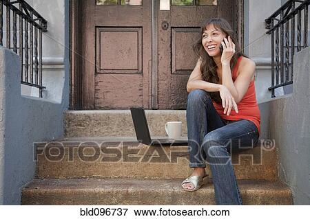 Stock Photo - Mixed race woman sitting on front stoop with cell phone and laptop. Fotosearch