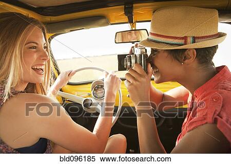 Stock Photography - Woman taking picture of friend driving old-fashioned car. Fotosearch