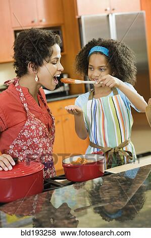 Stock Photo - African American daughter giving mother taste of sauce. Fotosearch