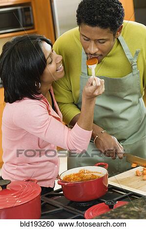 Stock Image - African American man tasking wife's sauce in kitchen. Fotosearch
