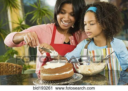 Stock Image - African American mother and daughter frosting cake. Fotosearch