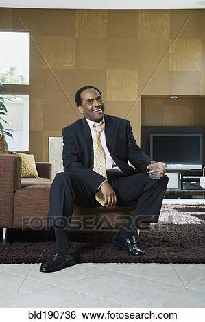Stock Photograph - African businessman sitting in modern living room with drink. Fotosearch