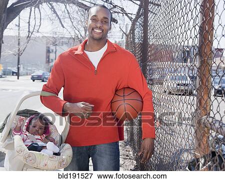 Stock Photo - African man carrying baby in carrier and holding basketball. Fotosearch