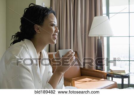 Stock Image - African woman drinking coffee in bedroom. Fotosearch