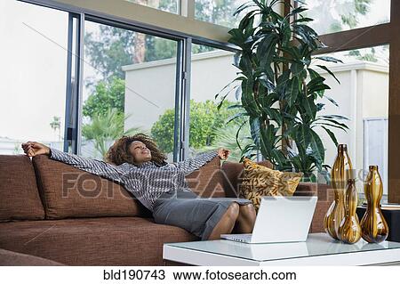 Stock Image - African woman relaxing on sofa in modern living room. Fotosearch