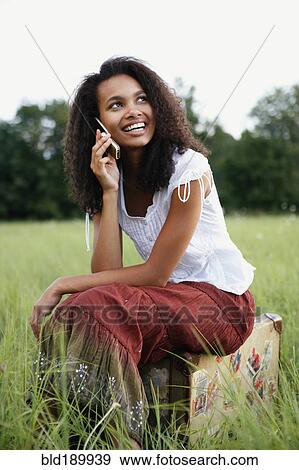 Stock Photo - African woman sitting on suitcase and using cell phone. Fotosearch