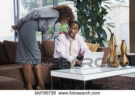 Stock Photo - African woman watching husband type on laptop in living room. Fotosearch
