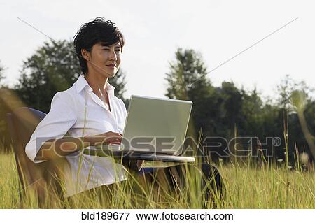 Stock Photo - Asian businesswoman typing on laptop in field. Fotosearch