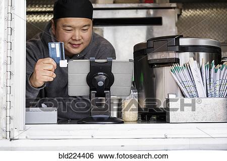Stock Photograph - Asian man processing credit card using digital tablet at food cart. Fotosearch