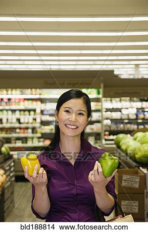 Picture - Asian woman deciding between green and yellow bell peppers. Fotosearch