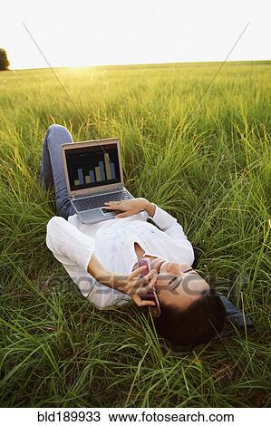 Stock Image - Asian woman laying in meadow using laptop and cell phone. Fotosearch