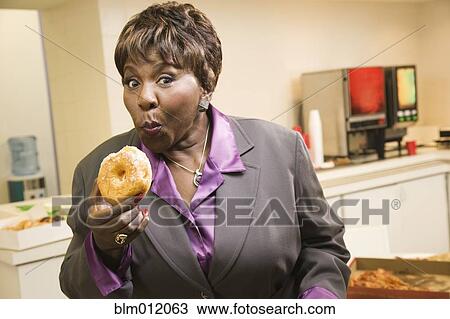 Stock Image - Black businesswoman eating donuts in office. Fotosearch