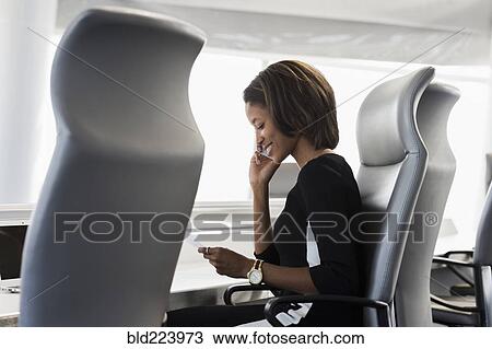 Stock Image - Black businesswoman reading paperwork talking on cell phone. Fotosearch