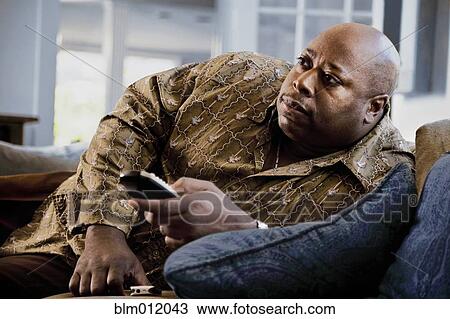 Stock Image - Black man reclining on sofa watching television. Fotosearch