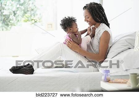 Stock Photo - Black woman cuddling with baby daughter on bed. Fotosearch