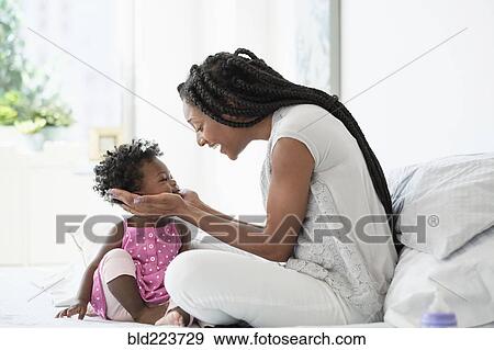 Stock Photo - Black woman cupping face of baby daughter on bed. Fotosearch