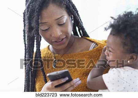 Stock Image - Black woman holding baby daughter using cell phone. Fotosearch