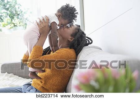 Picture - Black woman kissing nose of baby daughter on sofa. Fotosearch