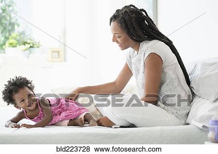 Stock Photo - Black woman playing with baby daughter on bed. Fotosearch