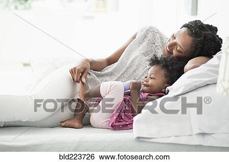 Stock Photograph - Black woman playing with foot of baby daughter on bed. Fotosearch
