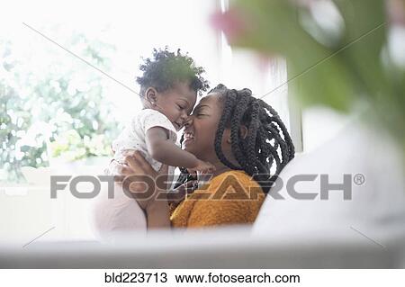 Stock Image - Black woman rubbing noses with baby daughter on sofa. Fotosearch