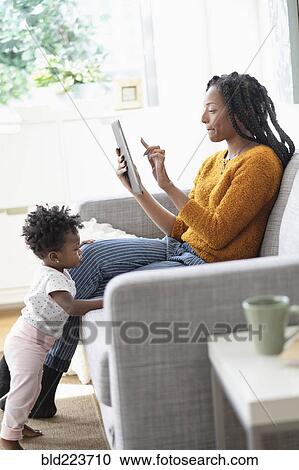 Stock Image - Black woman using digital tablet on sofa near baby daughter. Fotosearch