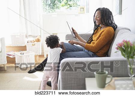 Stock Image - Black woman using digital tablet on sofa near baby daughter. Fotosearch
