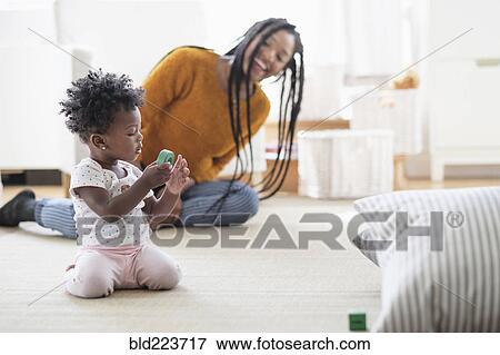 Stock Photo - Black woman watching baby daughter play with toy on carpet. Fotosearch