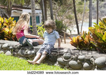 Stock Photo - Caucasian brother and sister sitting on stone wall at beach. Fotosearch