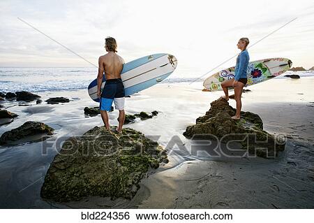 Stock Photograph - Caucasian couple standing on rocks carrying surfboards at beach. Fotosearch