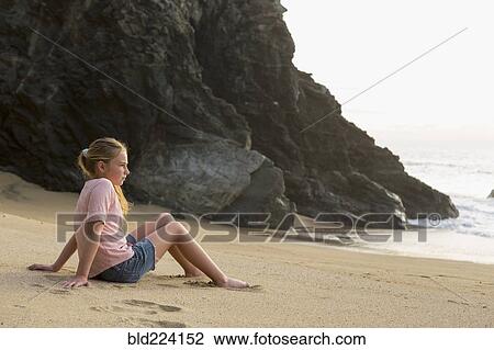 Stock Image - Caucasian girl sitting near rocks at beach. Fotosearch