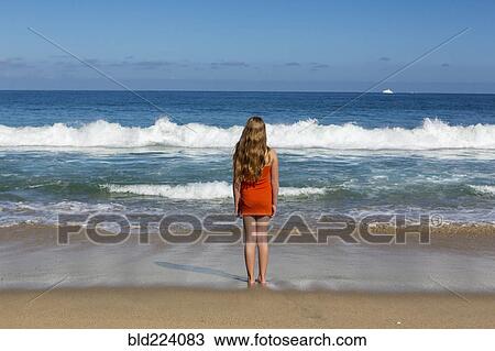 Stock Image - Caucasian girl standing looking at ocean waves. Fotosearch