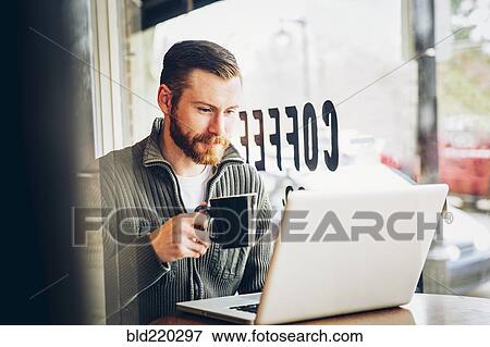 Stock Photo - Caucasian man drinking coffee and using laptop in cafe. Fotosearch