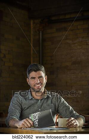 Stock Image - Caucasian man drinking coffee in cafe holding booklet. Fotosearch