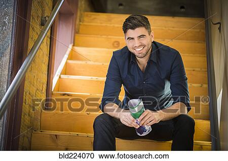 Stock Image - Caucasian man holding paperwork sitting on wooden staircase. Fotosearch