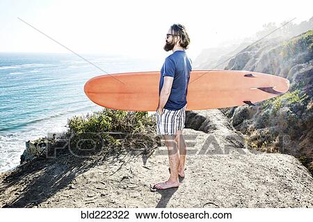 Stock Image - Caucasian man holding surfboard on coastal hilltop. Fotosearch