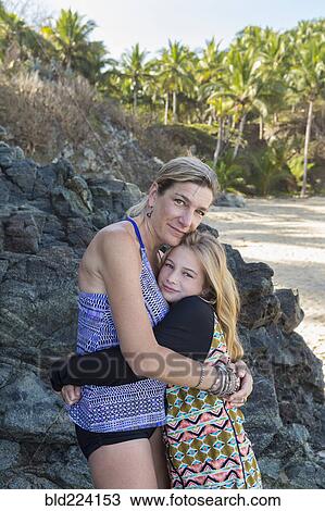 Stock Image - Caucasian mother and daughter hugging near rocks at beach. Fotosearch