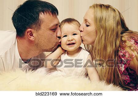 Stock Photo - Caucasian parents kissing baby daughter in bed. Fotosearch
