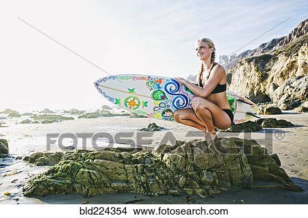 Caucasian woman crouching on rock carrying surfboard at beach View Large Photo Image Picture - Caucasian woman crouching on rock carrying surfboard at beach. Fotosearch