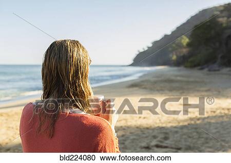 Stock Image - Caucasian woman drinking coffee on beach. Fotosearch