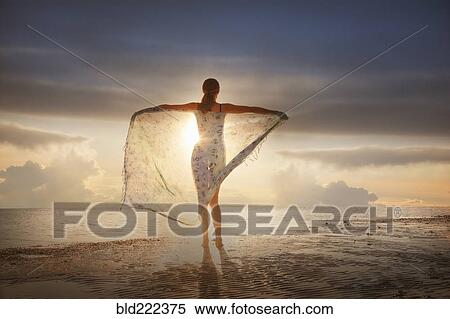 Stock Photography - Caucasian woman holding fabric on beach at sunset. Fotosearch